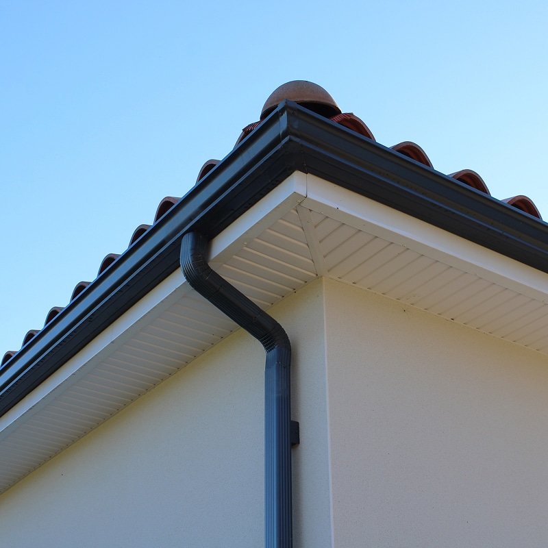 Gouttière noire et descente sur soffite blanc. Descente de gouttière noire fixée à l'angle d'une maison. Soffite blanc, mur en stuc clair et tuiles de toit.